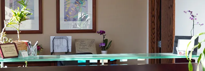 Reception desk with glass top, decorative plants, and framed artwork on the wall in a bright office.